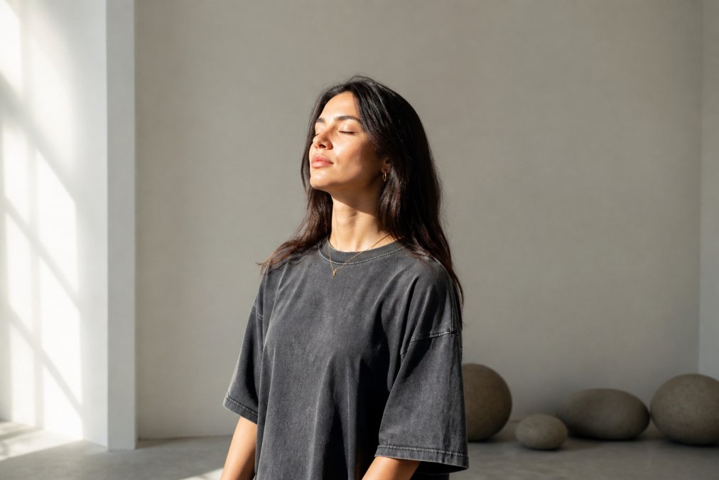 A young woman wearing an oversized vintage-washed graphic tee with clean lines, standing in soft natural light against a minimal neutral background, eyes closed in a calm, meditative pose