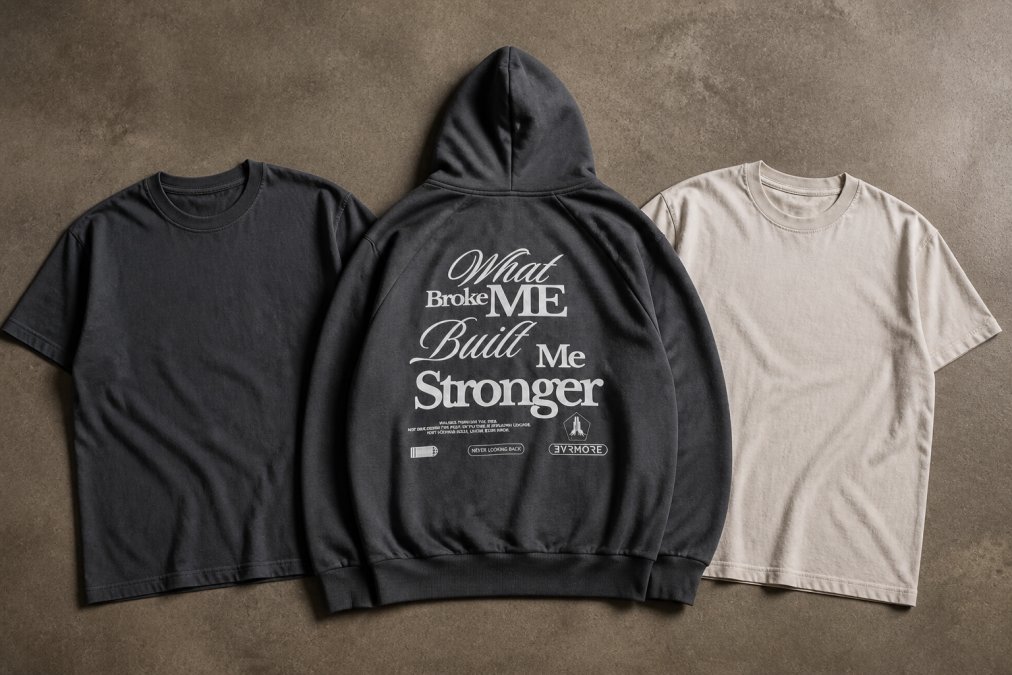 Flat lay of essential minimalist streetwear pieces arranged neatly in monochromatic colors on neutral surface, showing slate tee, black hoodie, and vintage pearl tee with quality fabric textures visible