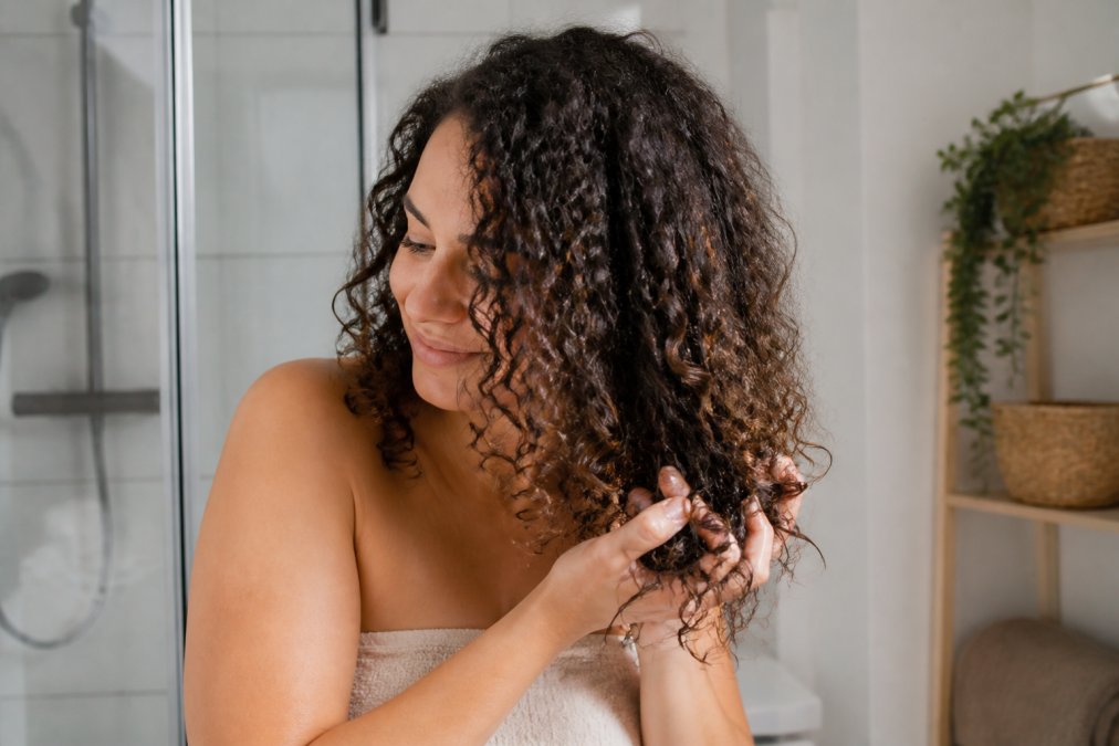 Vrouw met krullen knijpt stylingproduct in haar haar voor de curly girl routine in een lichte badkamer.