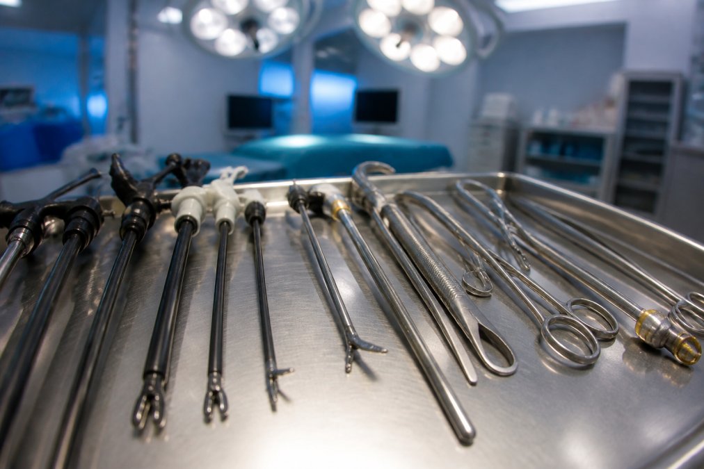 Close-up of surgical instruments arranged on a sterile metal tray in an operating theatre, showing delicate laparoscopic instruments with narrow shafts and articulated tips alongside traditional larger surgical instruments, with overhead operating lights reflecting off the metal surfaces