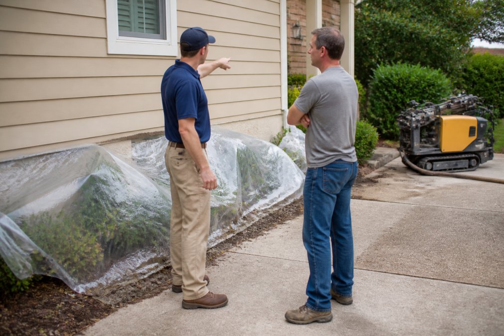 A homeowner in casual clothes standing in their driveway, talking with a foundation repair contractor who is pointing at the exterior wall of a house. Plastic sheeting covers nearby landscaping, and a small piece of equipment sits in the background. The scene is calm and professional, not chaotic.
