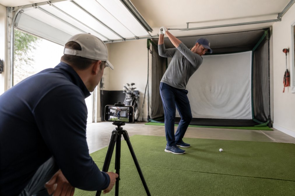 Golfer using a smartphone to check golf simulator ceiling height and swing clearance in a home garage setup.