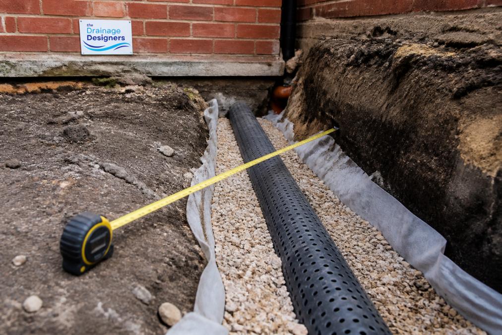Close-up of a construction site showing a perforated drainage pipe laid in a gravel trench near a brick building foundation, with a tape measure visible