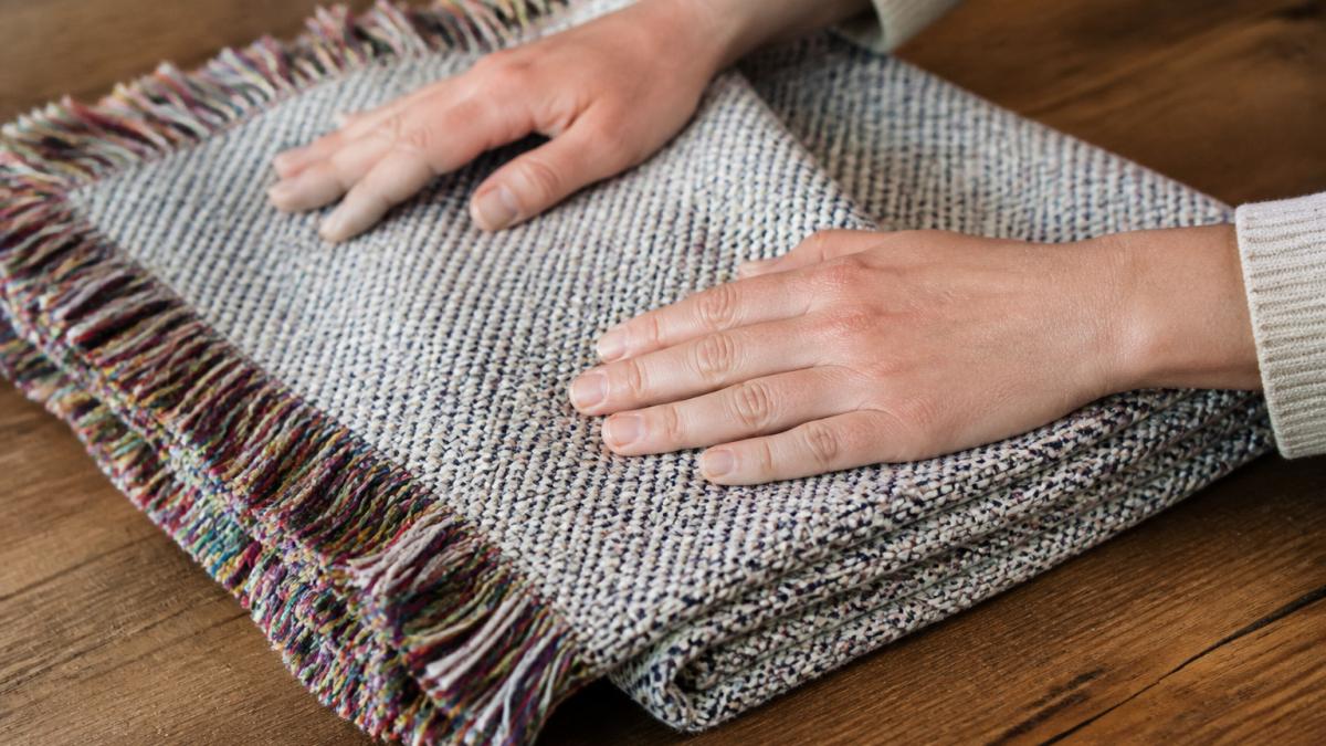 Close-up of hands touching a folded woven cotton blanket with colorful fringe on a wooden table, showing the texture of the weave