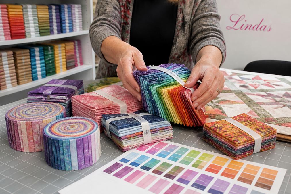 Quilter's hands arranging colorful precut fabric bundles including fat quarters, jelly rolls, and charm packs on a work table with a design layout and color swatches