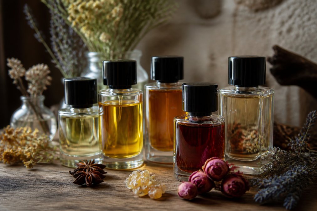 Close-up of fragrance bottles displaying different colored liquids, arranged on a wooden surface with dried flowers and botanical elements like lavender and vanilla pods nearby