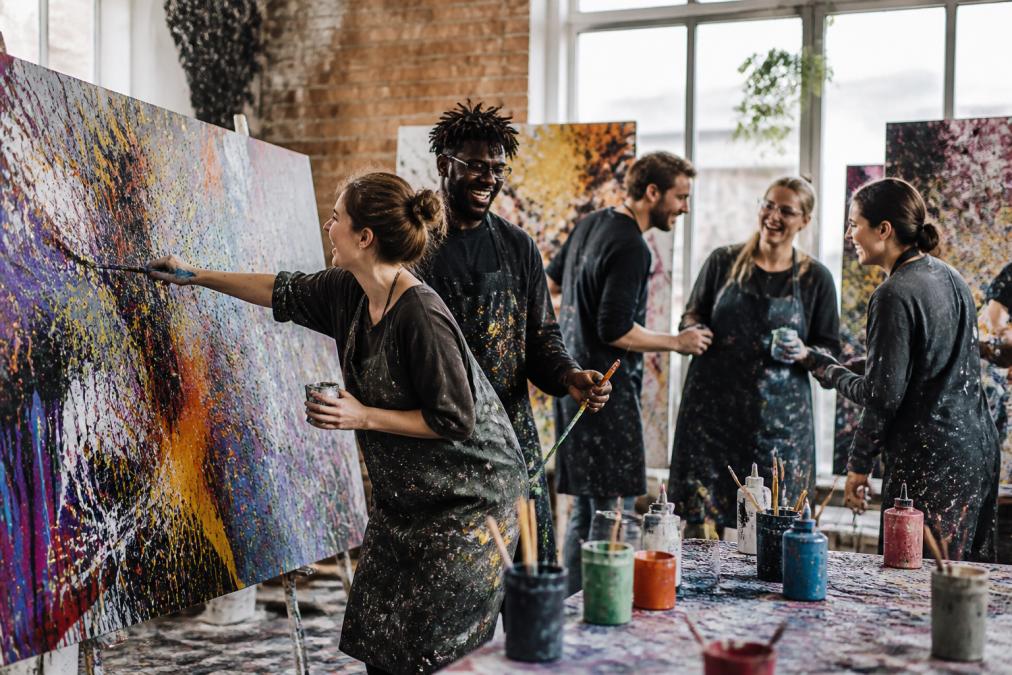Eine diverse Gruppe von Mitarbeitern malt gemeinsam auf großen Leinwänden, lacht und ermutigt sich gegenseitig während eines kreativen Action-Painting-Events in einem hellen, modernen Studio mit bunten Farben überall