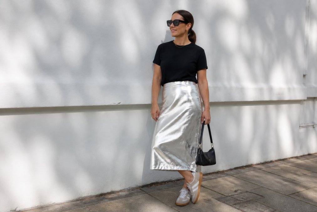 Woman wearing a silver metallic midi skirt paired with a plain black t-shirt and neutral trainers, standing in natural daylight against a white wall