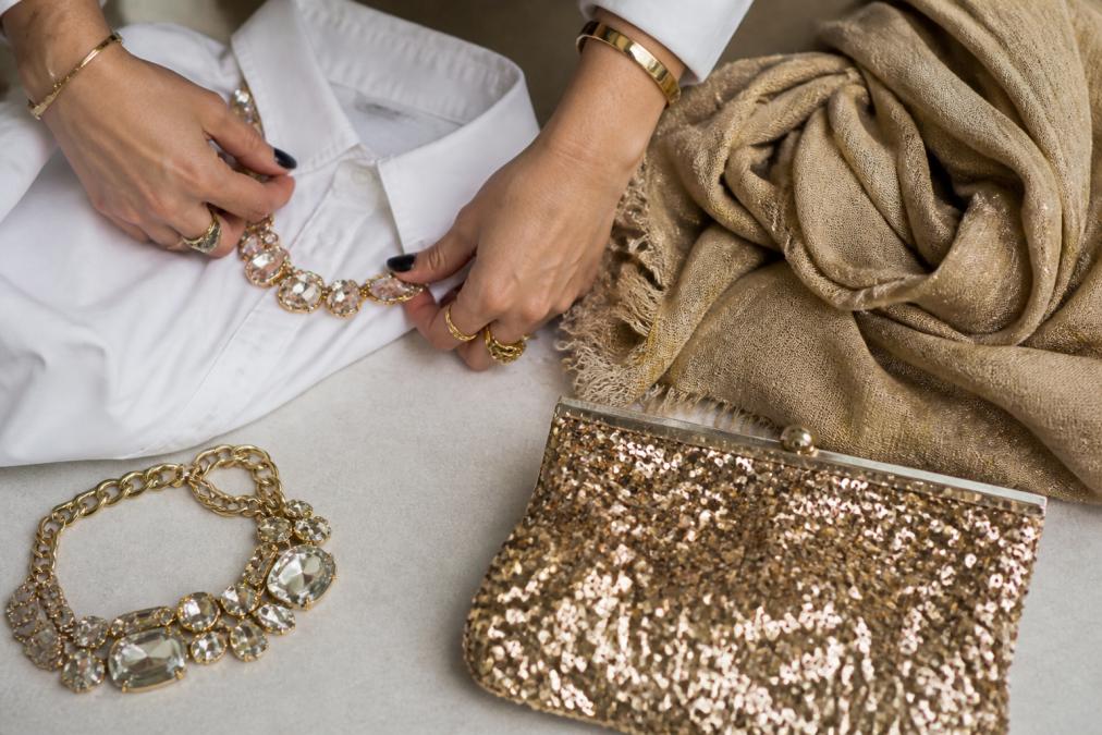 Close-up of hands styling a simple white button-up shirt with bold statement jewellery, a sequined clutch, and a textured scarf arranged on a neutral surface