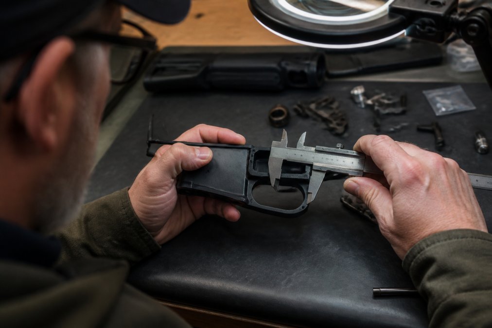 Detailed view of a gunsmith's hands inspecting an MKE lower receiver with precision calipers, measuring the trigger group pocket for tolerance stacking issues under bright magnification lighting
