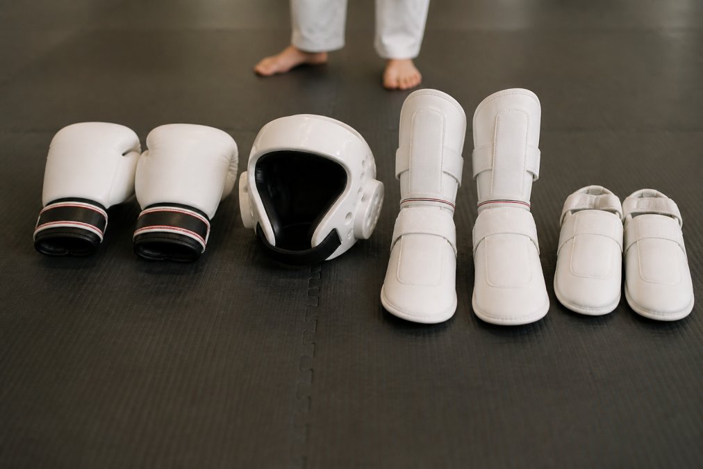 Flat lay photo of a complete competition sparring set including gloves, shin guards, headgear, and foot protectors arranged neatly on a dark gym mat, with an athlete's feet blurred in the background