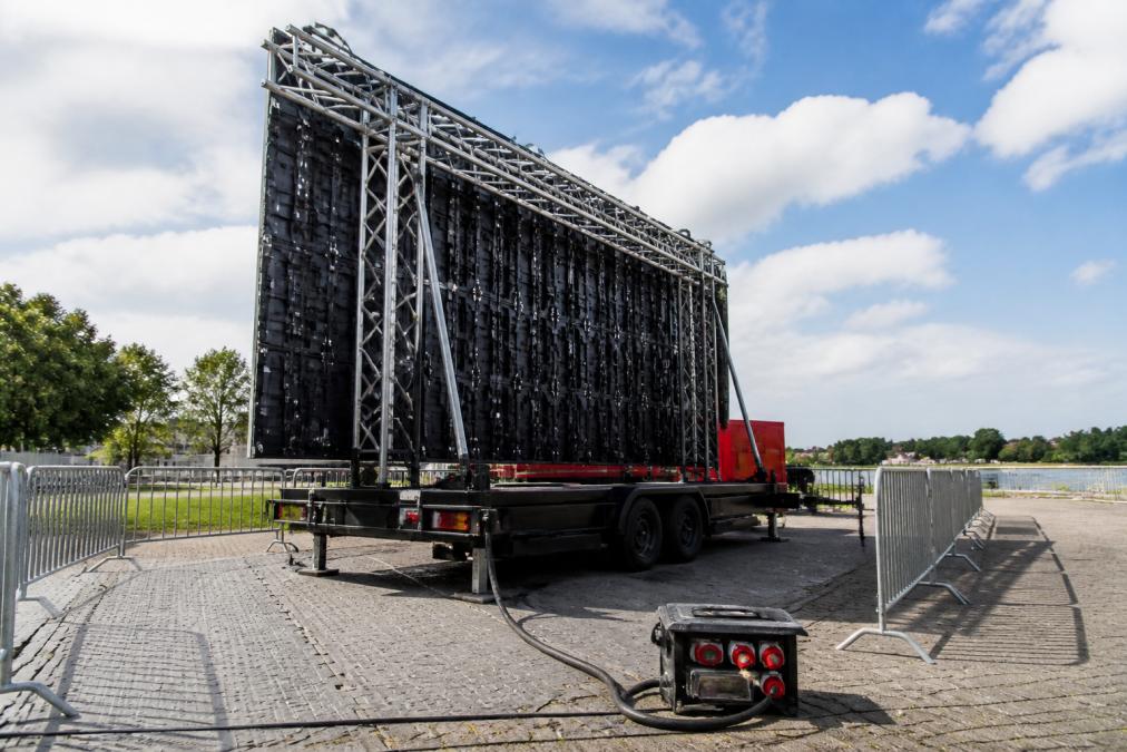 Mobiele LED-aanhangwagen volledig opgesteld op een evenemententerrein met zichtbare trussconstructie, aluminium steunbalken en stroomkabels in daglicht, technicus in veiligheidsvest voert controles uit
