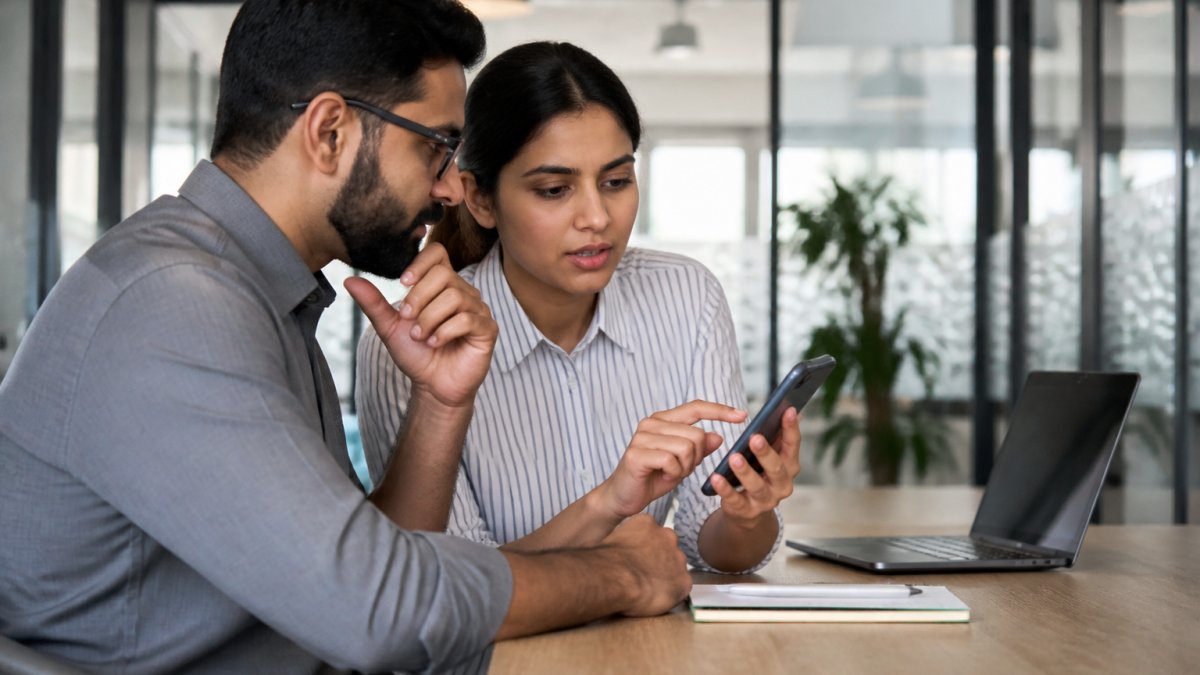 A developer and security tester reviewing a mobile app on a smartphone in a bright office, with a laptop showing test results beside them