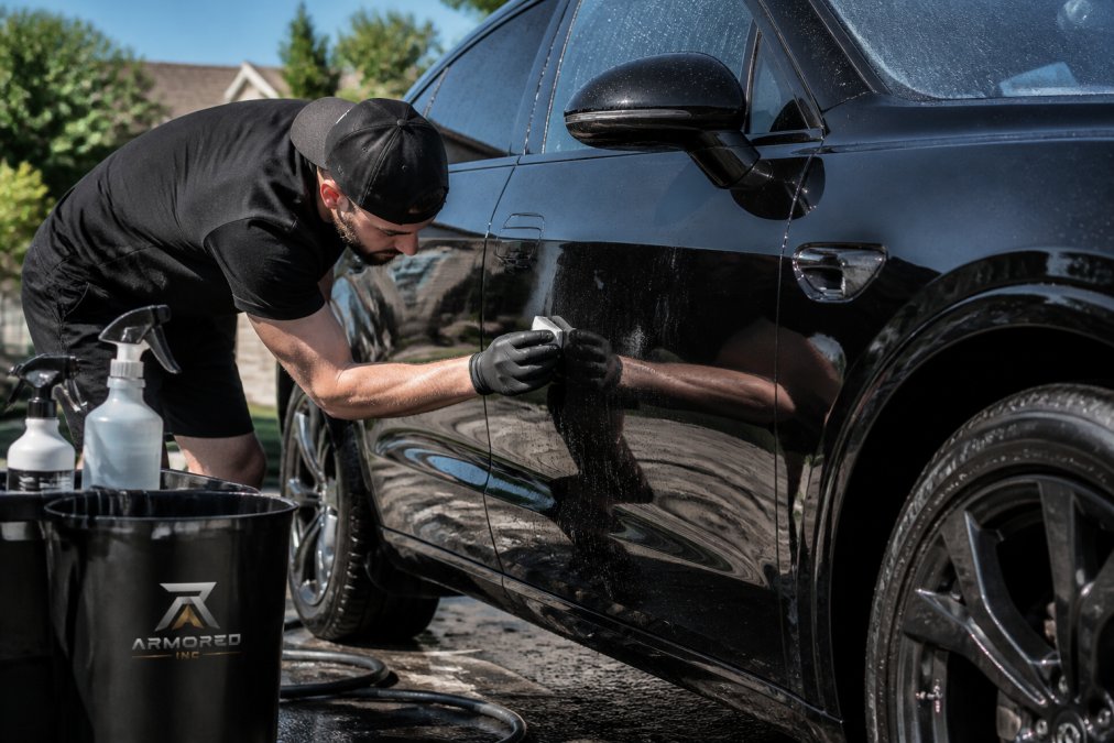 Professional detailer using clay bar treatment on a luxury vehicle's exterior, with water spray and detailing supplies visible, showing precision hand technique on a sunny driveway