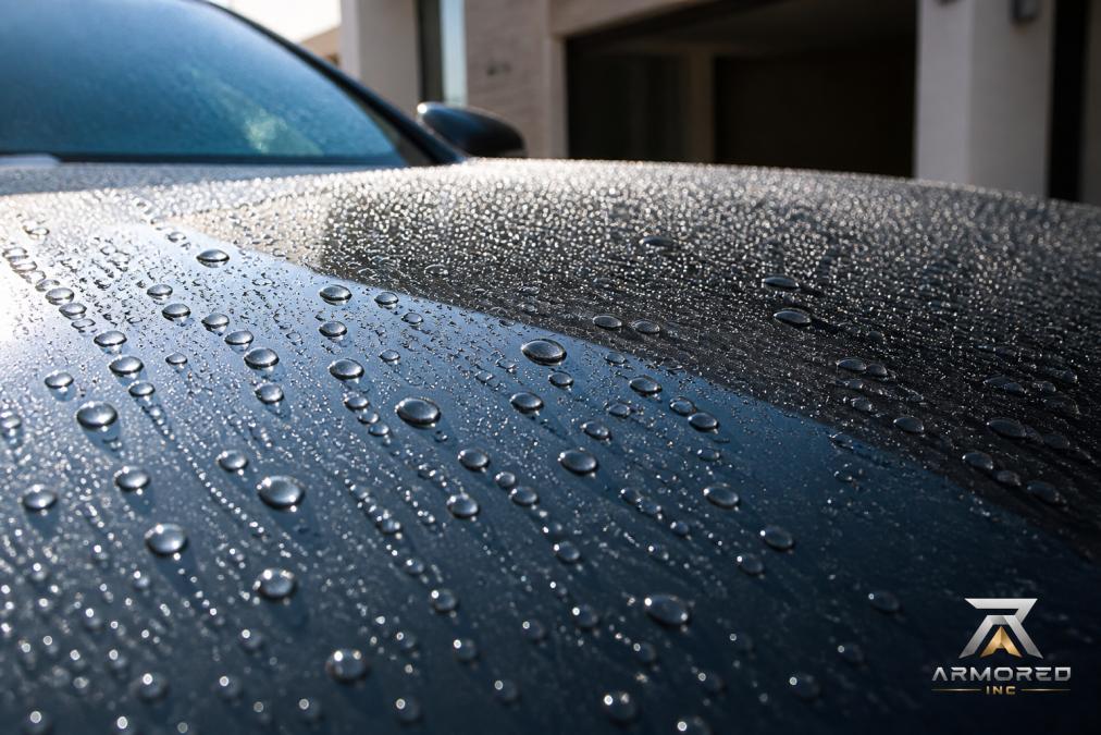Close-up of water beading on a freshly ceramic-coated car hood, showing hydrophobic surface with water droplets rolling off in bright sunlight