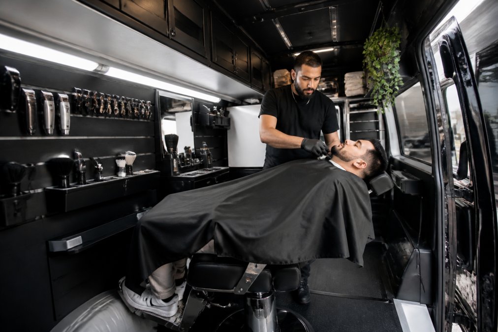 Professional barber working at a compact mobile barber station inside a van with a reclining barber chair, wall-mounted clipper storage, LED lighting strips, and organized tool trays in a cleanly organized interior space