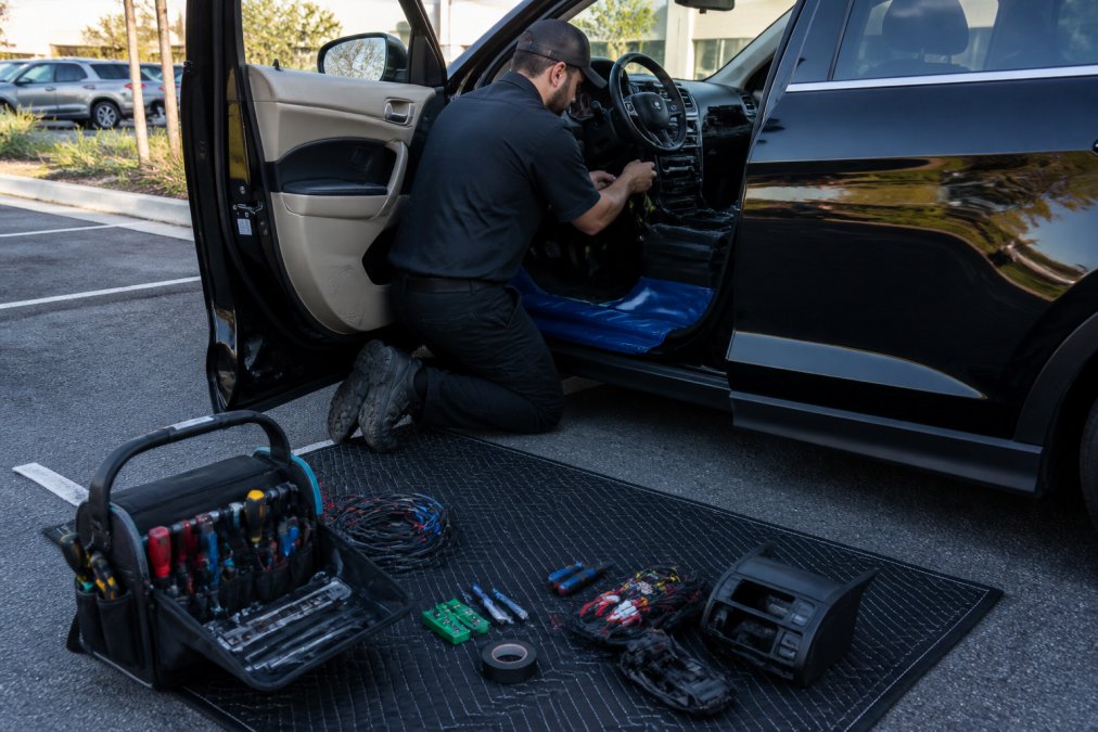 A mobile technician in a clean uniform working on a car dashboard in an office parking lot, tools organized on a mat, driver's door open, daylight