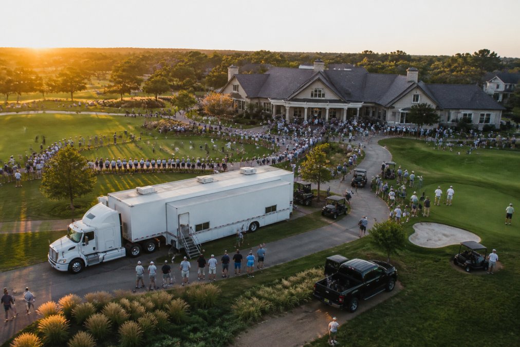 Aerial view of a sprawling golf tournament venue at golden hour, with a high-roof mobile command trailer positioned strategically near the clubhouse and first tee, staff moving between the unit and the course