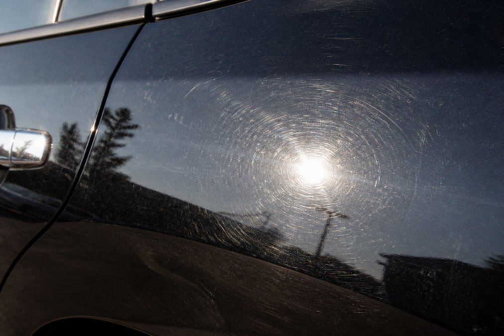 Close-up of a vehicle's paint surface showing swirl marks and water spots after automatic car wash, with harsh lighting highlighting the imperfections on the clear coat
