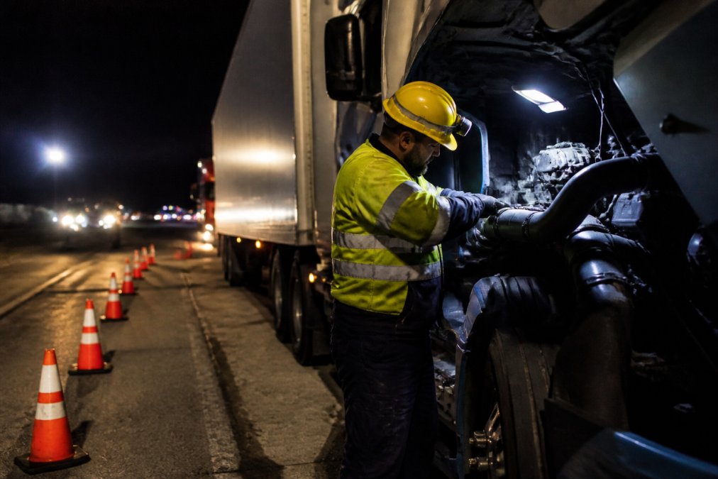 A mobile diesel mechanic working safely on the side of a highway at night with proper LED work lighting, orange safety cones positioned behind the truck, and the technician wearing high-visibility reflective clothing and a hard hat while working on a semi truck engine