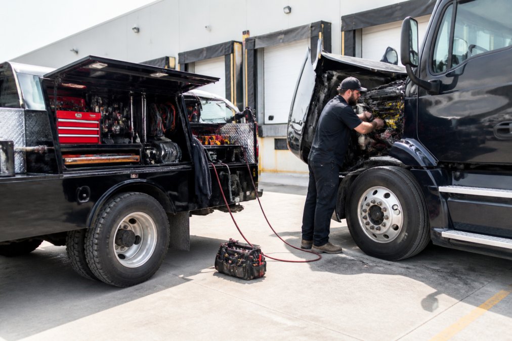 Fully equipped mobile service truck parked at a commercial loading dock with technician performing electrical diagnostics on a heavy-duty semi-truck, tools organized on the truck bed, bright daylight illuminating the work area