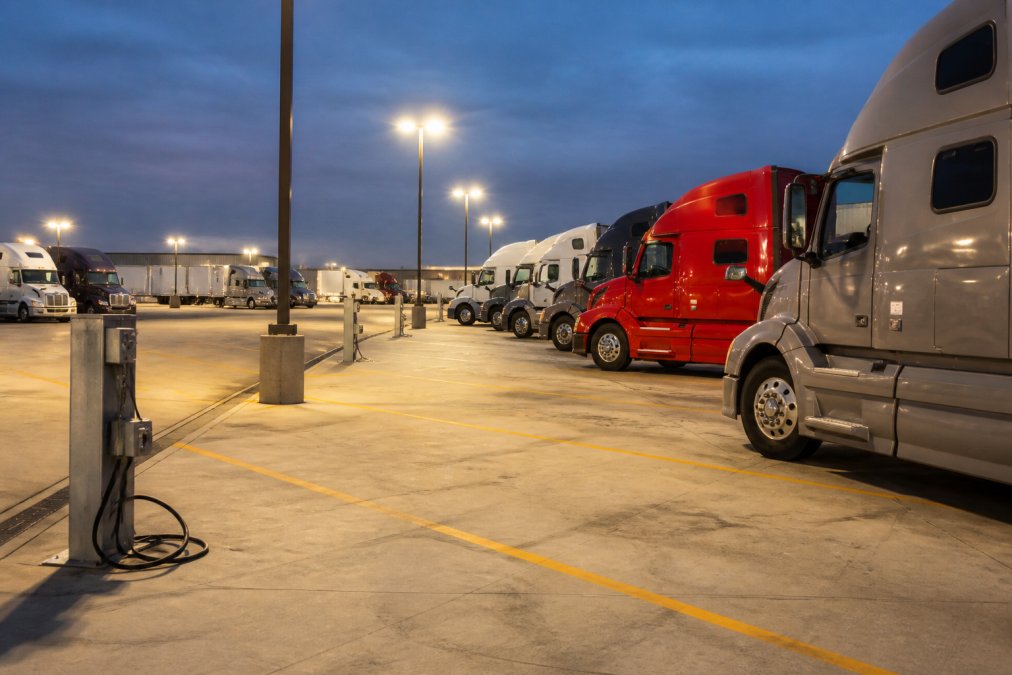Clean, well-lit yard terminal with multiple semi-trucks lined up in organized rows, showing adequate spacing between vehicles, bright overhead lighting illuminating the work area, and clear ground surface with power access points visible