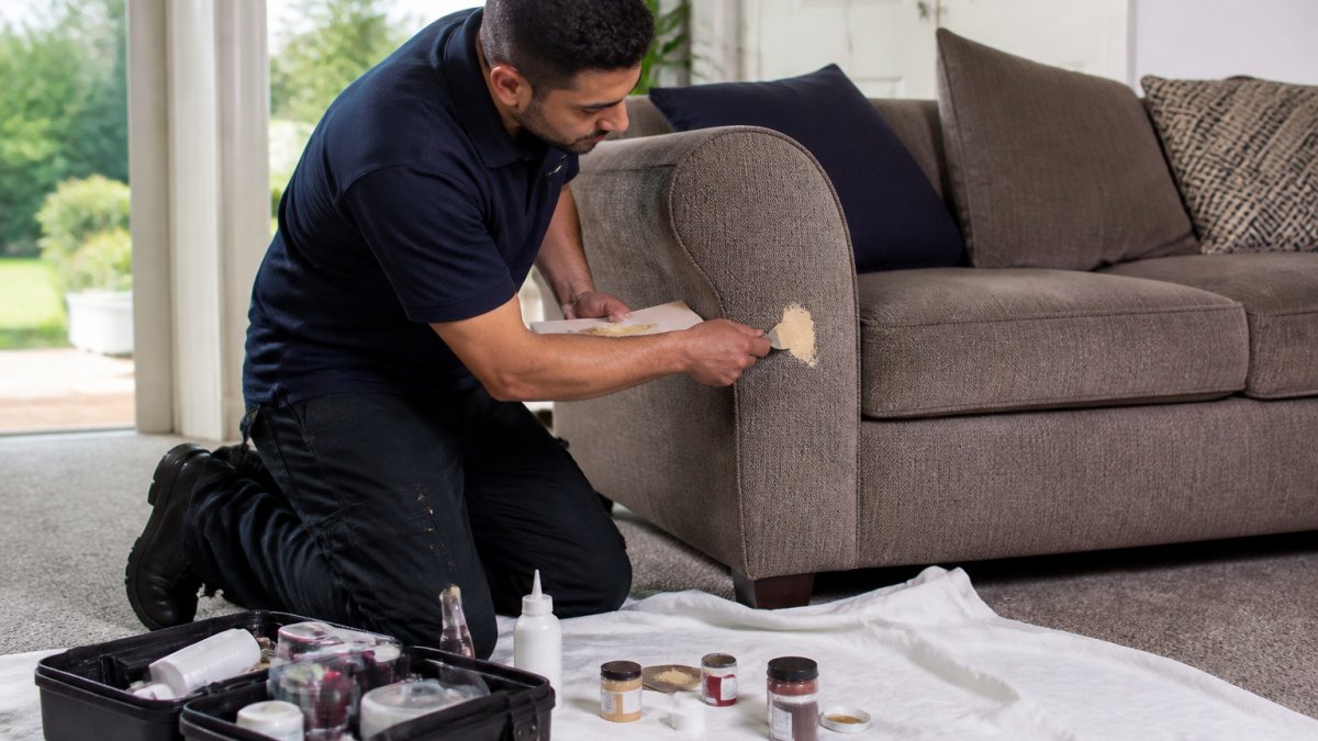 A furniture repair technician in a polo shirt kneeling beside a fabric sofa in a bright living room, applying colour-matched filler to a damaged armrest with a small tool, with a protective sheet on the carpet