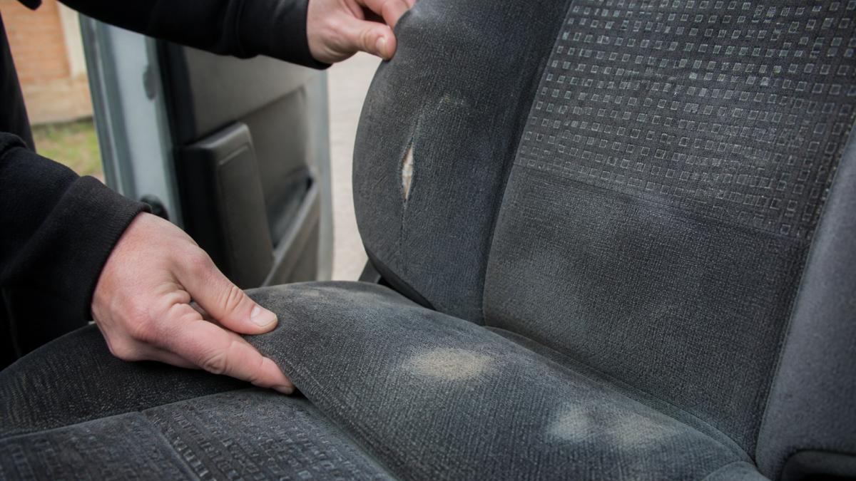 Close-up of a technician's hands examining a worn van seat fabric, showing visible scuff marks, discolouration patches, and a small tear in velour upholstery, with natural daylight illuminating the damage detail
