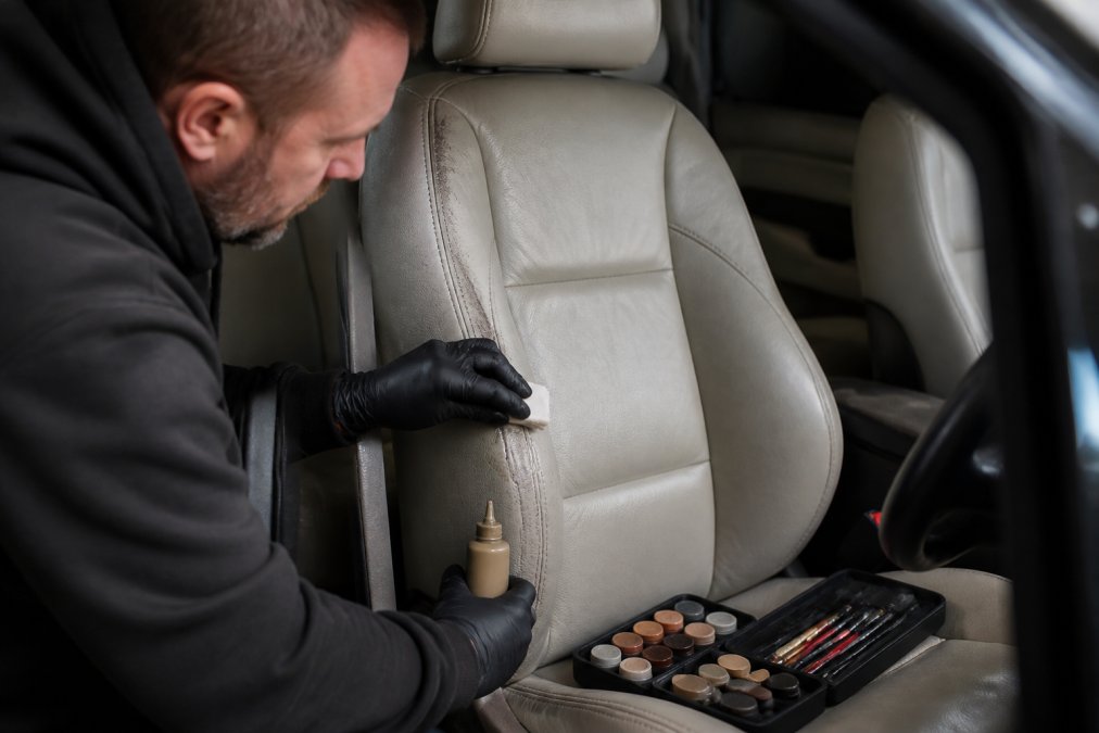 Professional demonstrating mobile leather seat repair cost technique on car seat with visible scuffs, using colour matching tools and applicators under bright workshop lighting