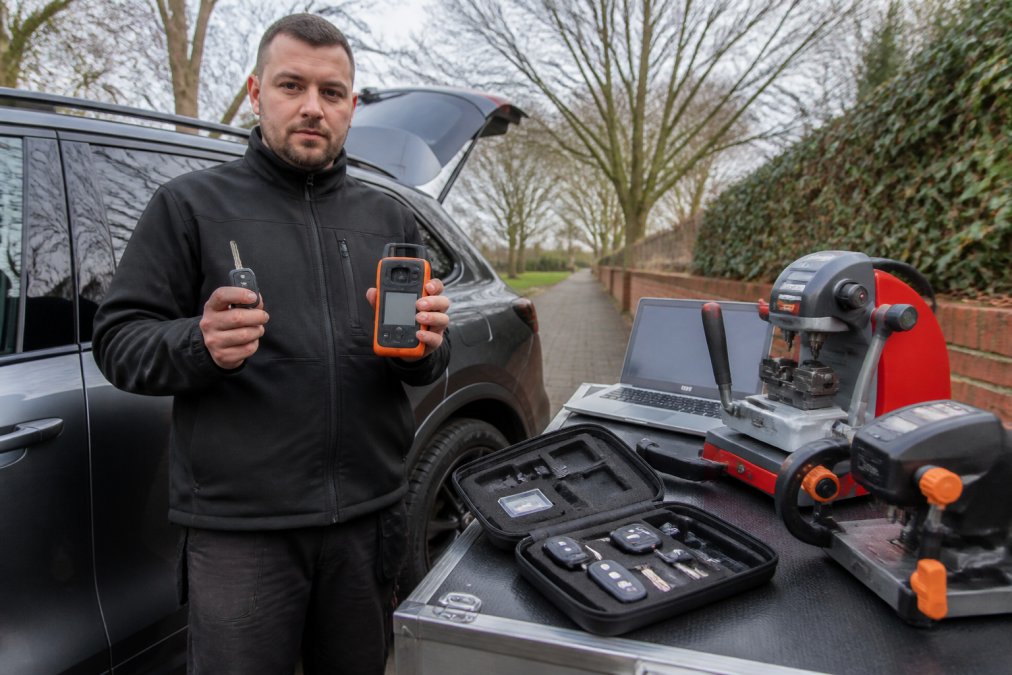 Professional mobile locksmith technician standing beside a vehicle with diagnostic equipment and laptop, holding a replacement transponder key and programming device, demonstrating on-site key cutting setup in natural daylight