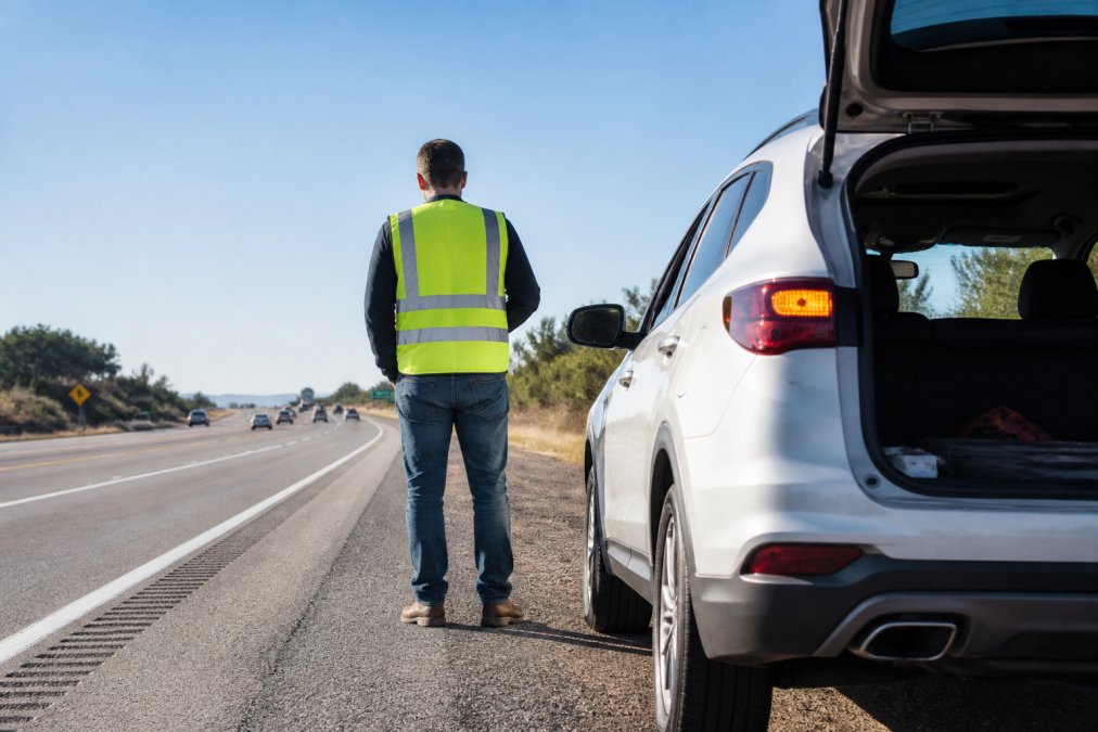 Driver standing safely behind vehicle on highway shoulder with hazard lights flashing, wearing reflective safety vest, daytime conditions