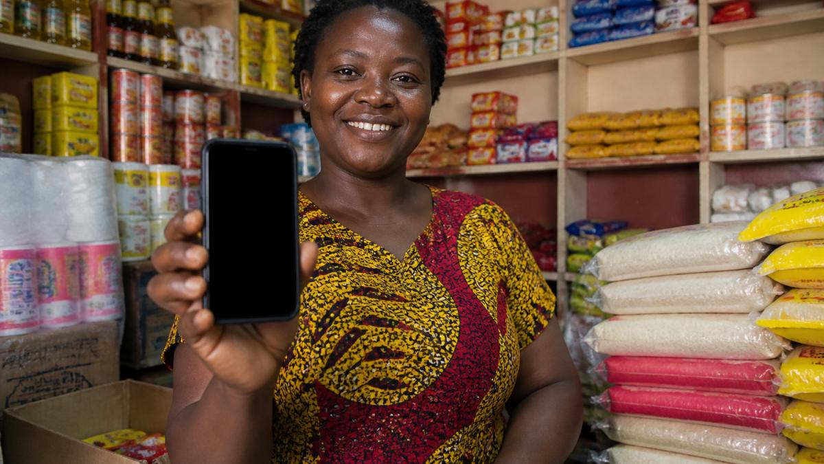 A Ghanaian shop owner holding a smartphone showing a mobile money payment confirmation screen, with shelves of bulk goods visible in the background