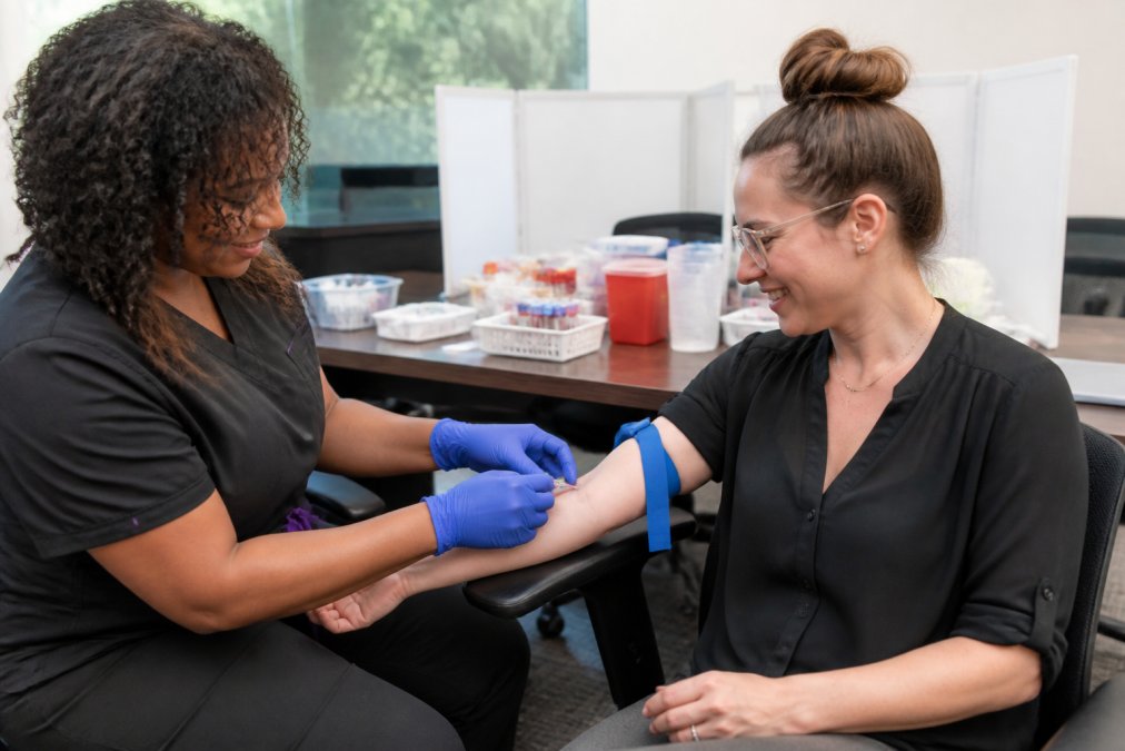 Licensed phlebotomist drawing blood from an employee seated comfortably in an office conference room, with biometric screening equipment and privacy screens visible on nearby tables