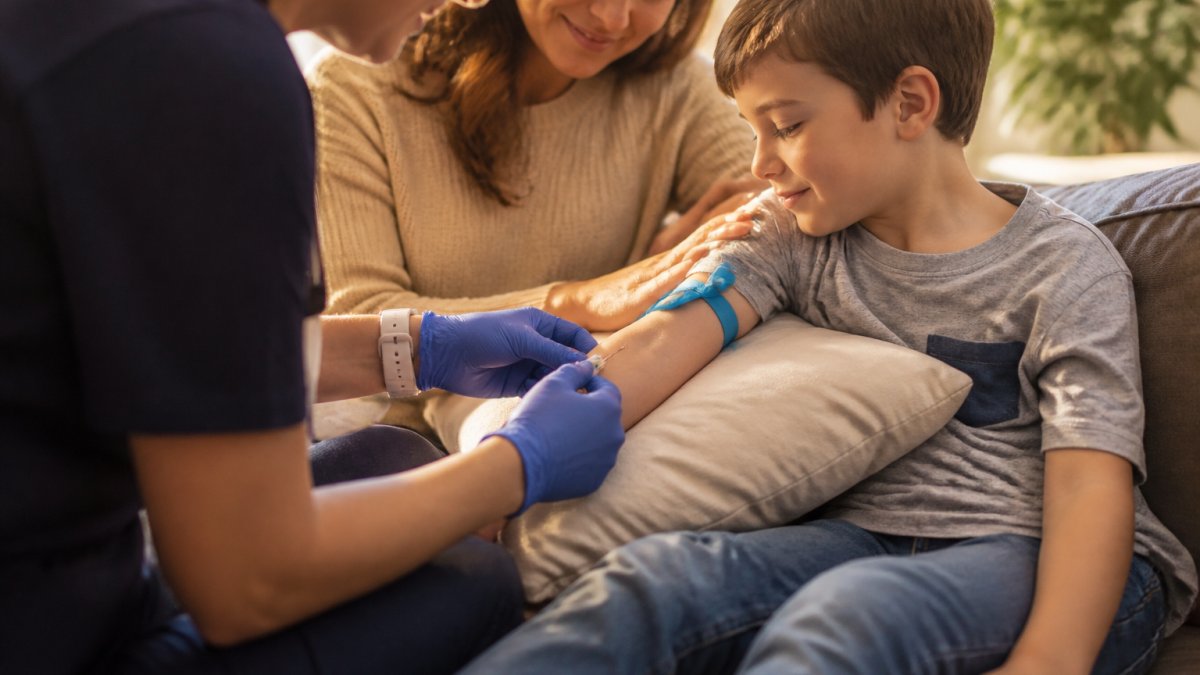 Professional phlebotomist performing a gentle blood draw on a young child's arm in a comfortable home setting, with a parent seated nearby providing reassurance and comfort