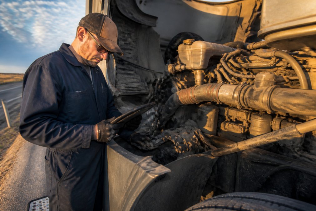 A mobile diesel mechanic in work uniform reviewing a heavy-duty semi-truck engine bay on the shoulder of a highway, diagnostic tablet in hand, afternoon sunlight casting long shadows across the truck's frame