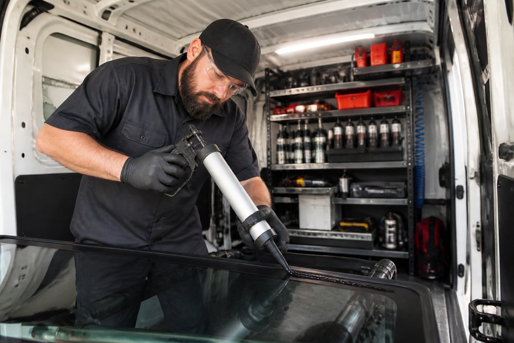 A certified technician in a branded uniform applying urethane adhesive to a new windshield inside a clean, well-lit service van with organized tools and calibration equipment on shelves