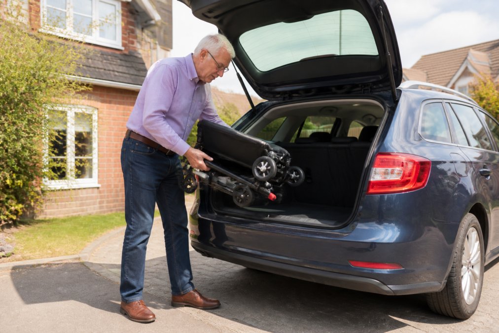 A person with limited mobility pressing a button on a remote control to fold a lightweight electric wheelchair, then lifting it into the boot of a small family car in a residential driveway on a sunny day