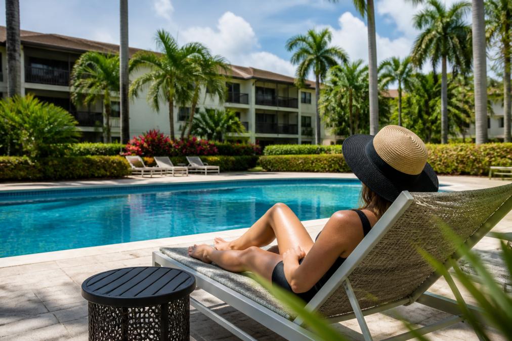 Modern Apartments Near Tri Rail in Boca Raton 2 Resident relaxing on a lounge chair by the resort-style pool surrounded by lush, manicured green spaces, palm trees, and flowering landscaping on a bright afternoon