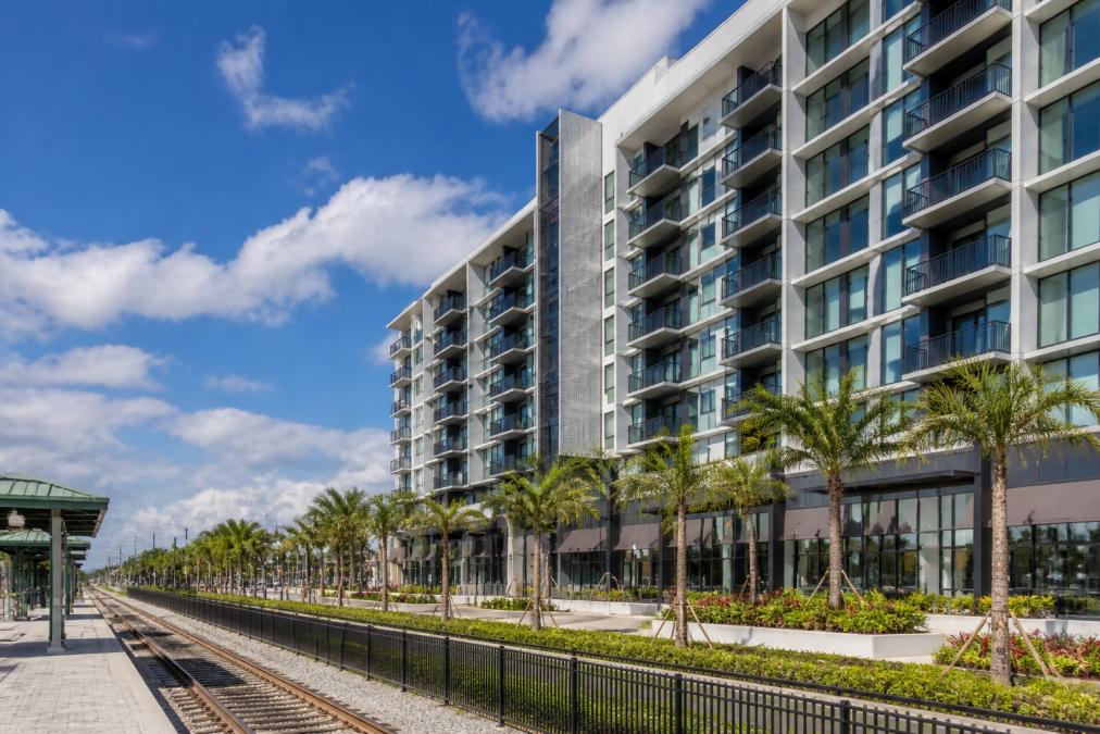 Modern Apartments Near Tri Rail in Boca Raton 3 Modern 8-story mixed-use apartment building with contemporary glass and metal facade, ground-floor retail storefronts with large windows, and landscaped plaza area on a clear day