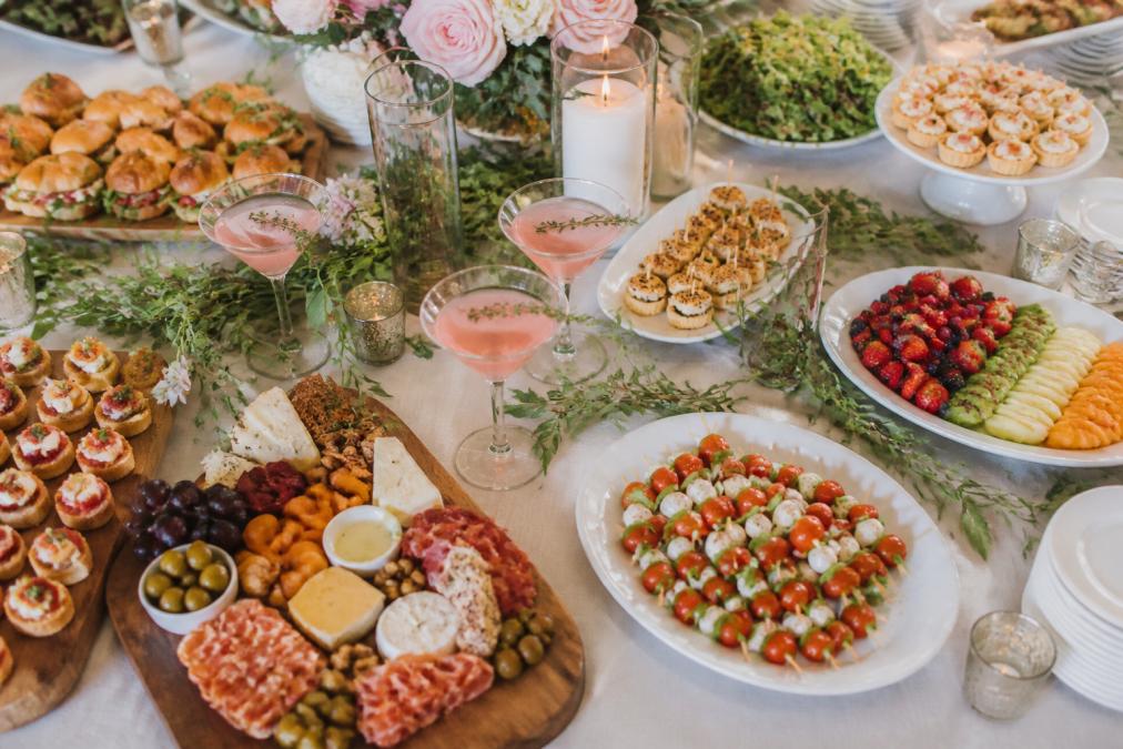 Overhead view of an elegant bridal shower spread with charcuterie boards, fresh fruit platters, finger foods, and signature cocktails arranged on a decorated table