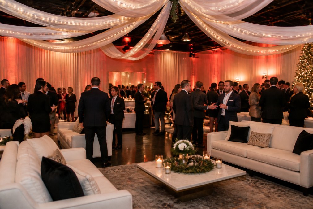 Elegantly decorated modern event space with contemporary lounge furniture, ambient up-lighting in warm tones, and draped fabric accents, with professional guests mingling during a corporate holiday celebration in a sophisticated atmosphere