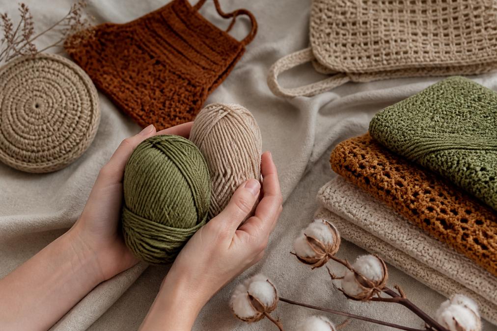 Hands holding natural fiber yarn in eco-friendly materials, with finished crochet pieces in muted, sustainable color tones arranged on natural linen fabric