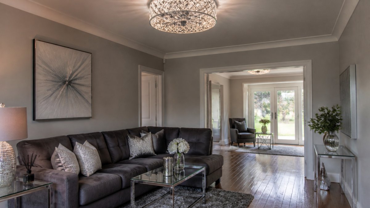 Modern living room with soft grey walls and wooden flooring, featuring an elegant flush-mount crystal ceiling light with a geometric ring design in polished chrome, creating warm ambient lighting that reflects off crystal accents