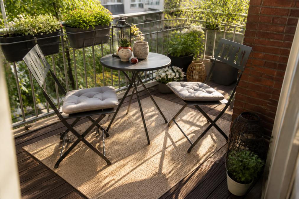 A cosy balcony setup featuring a small round bistro table with two folding chairs, a neutral-toned weather-resistant outdoor rug anchoring the seating area, potted plants arranged along the railings, and warm afternoon light creating welcoming shadows across the space