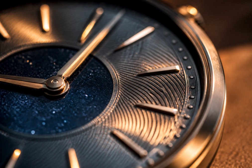 Macro photograph of a watch dial with intricate guilloché pattern, applied indices catching warm side light, deep blue and starlit texture visible in the center