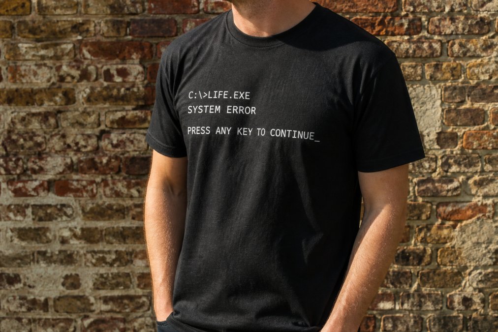 Close-up of a person's torso wearing a black graphic t-shirt with bold white retro computer text, standing against a weathered brick wall in natural daylight