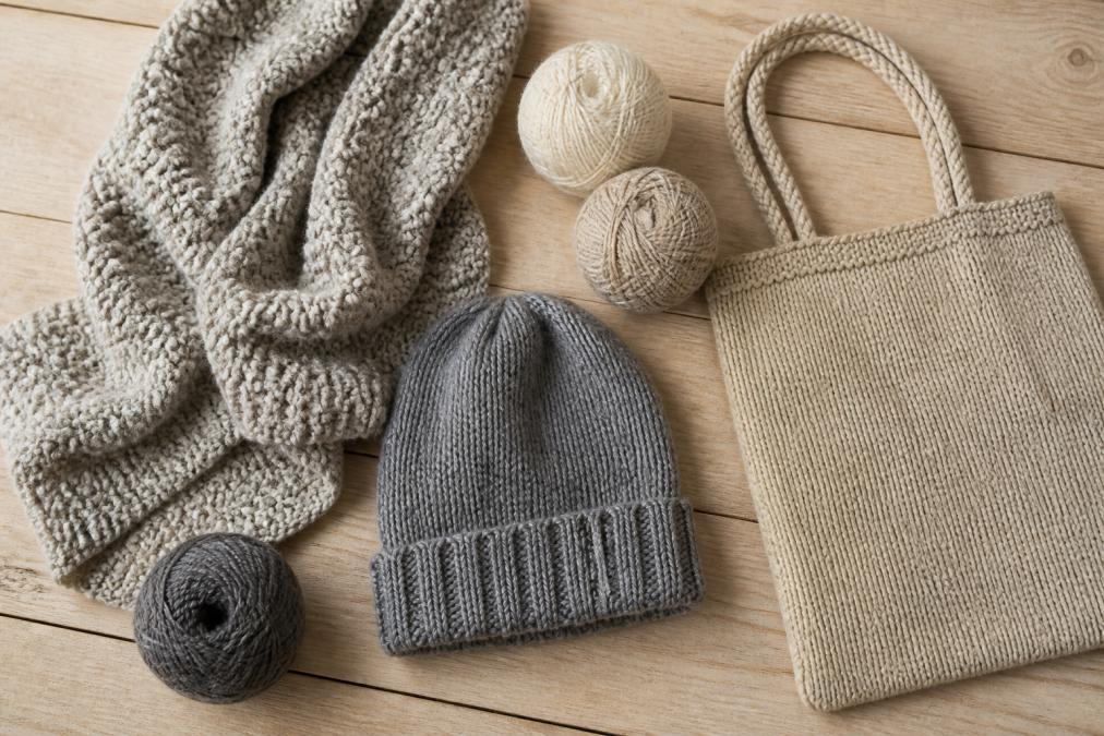 Flat lay of hand-knitted items including a chunky textured scarf, a ribbed grey beanie, and a simple rope-handled tote bag arranged on a light wooden surface with balls of neutral wool in cream, oatmeal, and slate