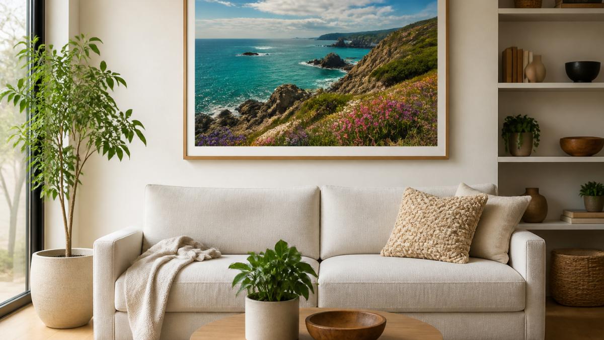 A bright modern living room with a neutral sofa and a large framed colourful landscape print centred above it, soft daylight from a side window, styled with a plant and a textured cushion