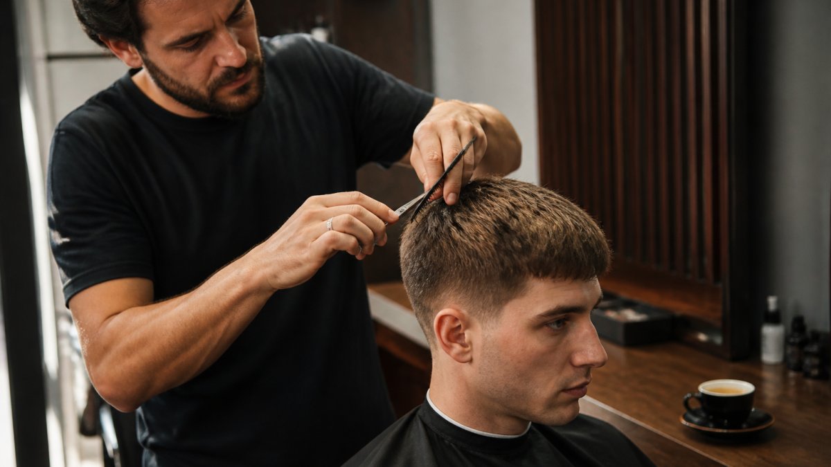 A barber using scissors and a comb to add texture to a men's short crop haircut in a relaxed studio, with warm lighting and an espresso cup on the counter nearby