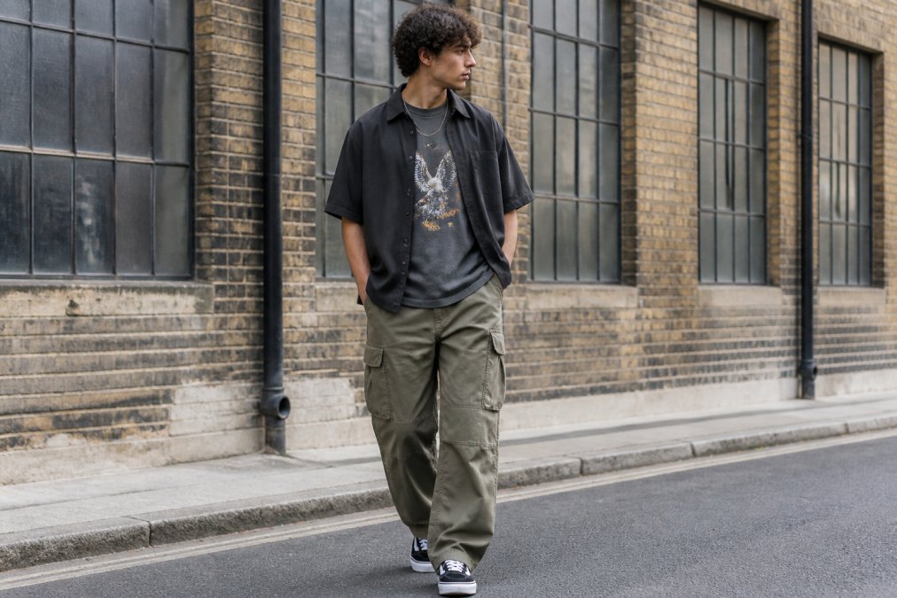 Male model wearing a layered streetwear outfit with a graphic tee visible under an open overshirt and baggy cargo pants, photographed on a city street in natural overcast light